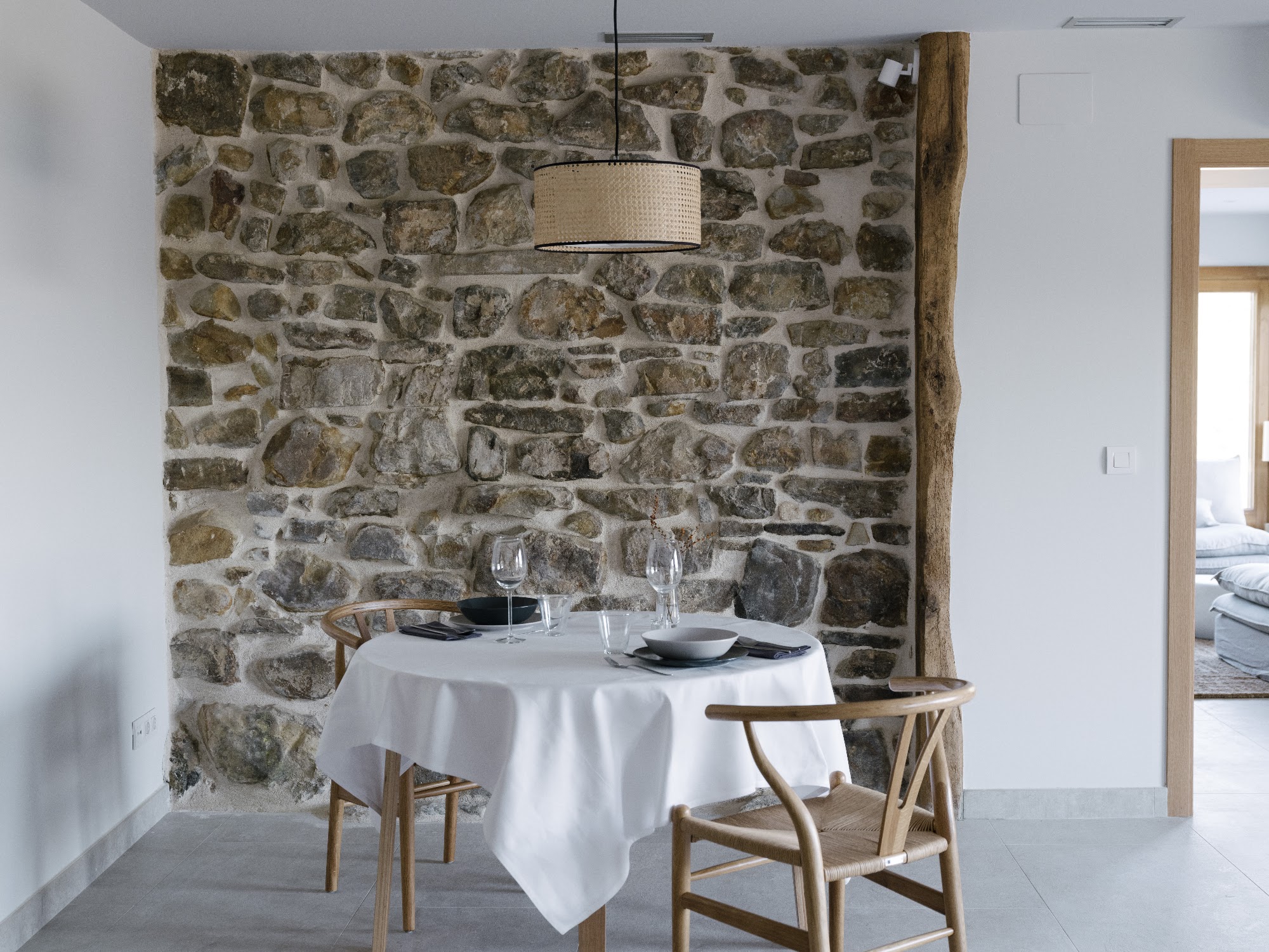 Comedor de ROLA: mantel blanco, sillas de madera y muro de piedra vista